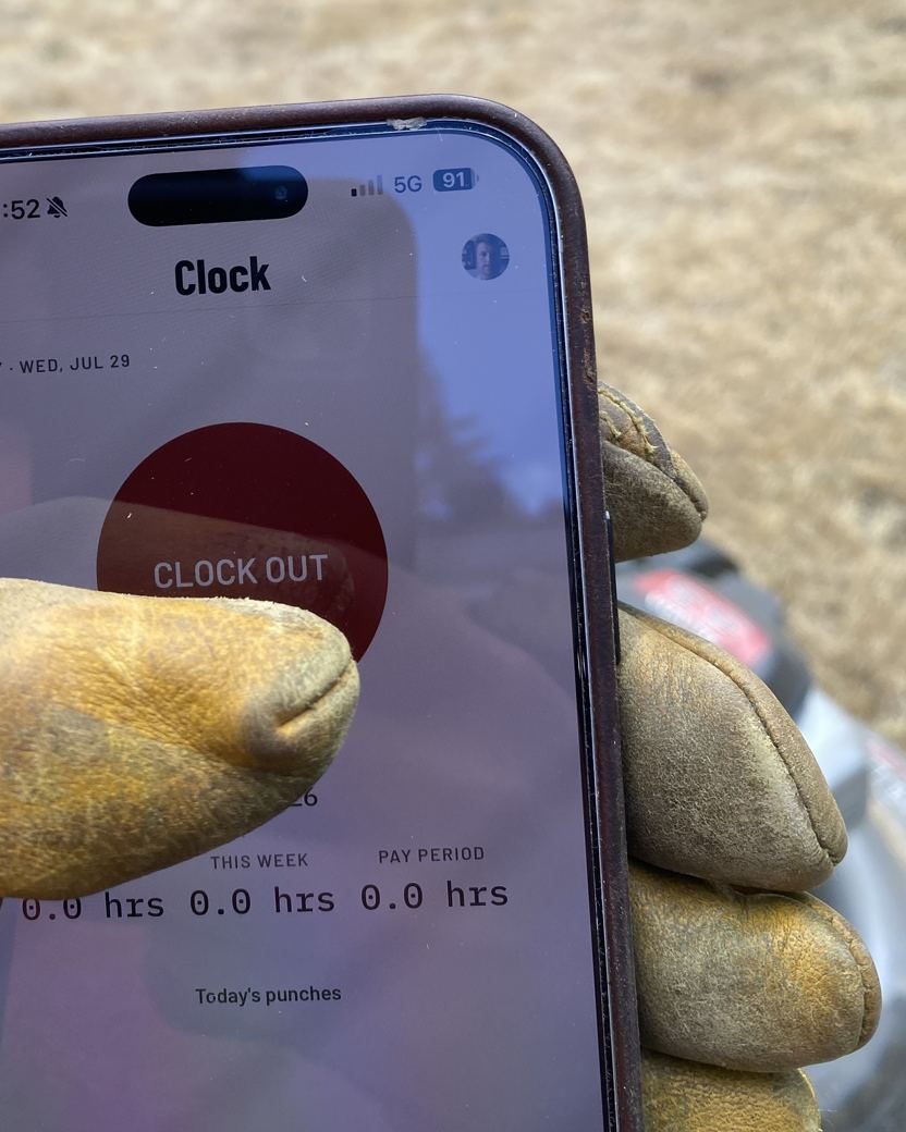 A work-gloved hand holding a phone running CutCrew’s Clock screen mid-job: a red CLOCK OUT button above the day’s hours.