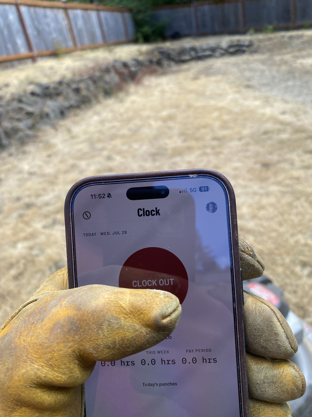A work-gloved hand holding a phone running CutCrew&rsquo;s Clock screen &mdash; a red CLOCK OUT button above the day&rsquo;s hours &mdash; standing over a freshly cut yard.